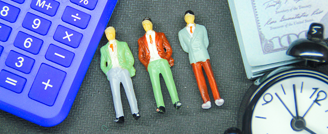Three figurines of business people in suits laying on a table. Its cropped with a calculator, money and a clock around the perimeter.